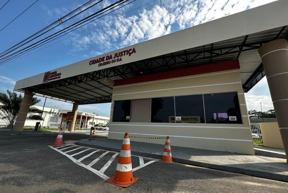 Foto da entrada da Cidade da Justiça de Cruzeiro do Sul. Mostrando a guarita, cones de transito na frente. A guarita é coberta por um grande telhado onde está a logomarca do Poder Judiciário e está escrito "Cidade da Justiça: Cruzeiro do Sul", tudo na cor vermelho escuro