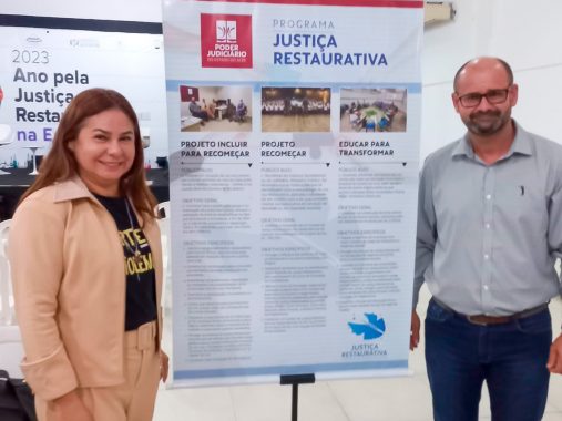 Fotografia da desembargadora Waldirene Cordeiro e do servidor Fredson Pinheiro ao lado do banner da Justiça Restaurativa, que possui informações dos projetos em andamento no Acre