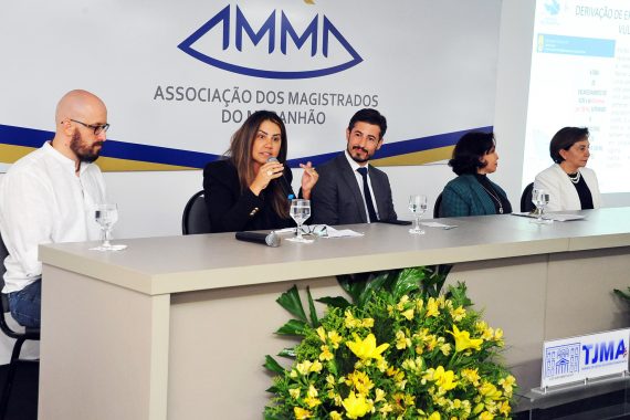 Fotografia da bancada de debatadores do painel "Possibilidades e Práticas nos Sistemas Criminais e Socioeducativo" e a juíza Andrea Brito está falando ao microfone