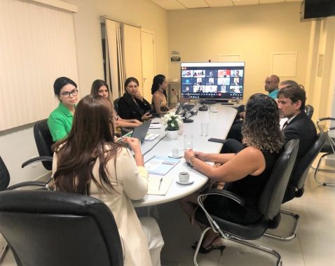 Fotografia da reunião híbrida do Comitê de Políticas Penais. Há participantes sentados à mesa e uma tela com participantes em videoconferência