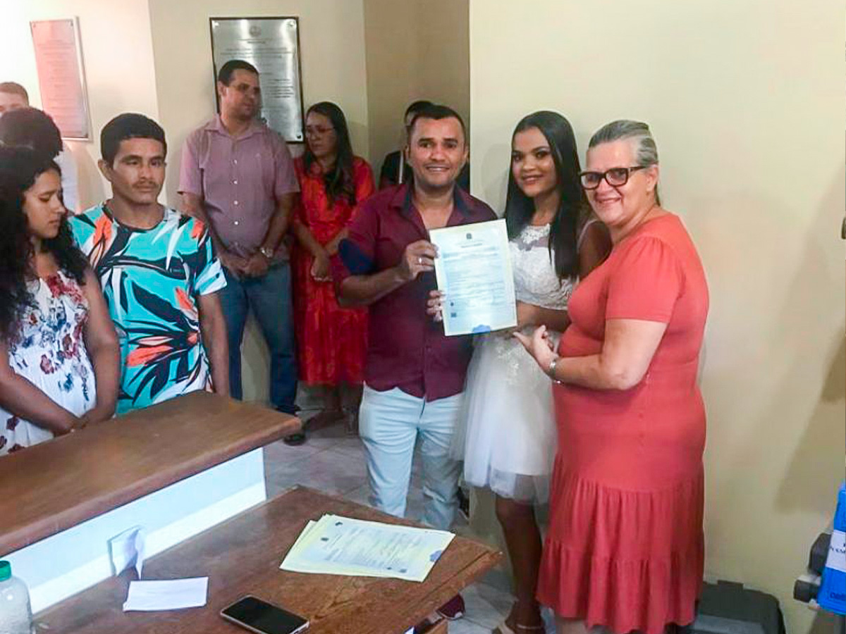 Servidora do TJAC está ao lado de um casal que participou do casamento coletivo. Todos estão mostrando juntos a foto da Certidão de Casamento