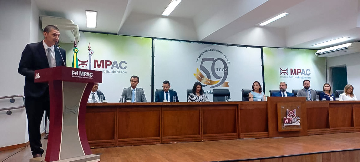 Fotografia do novo superintendente regional do trabalho falando no púpito na solenidade de sua posse. Atrás está uma bancada de autoridades que prestigiaram o evento no auditório do MPAC