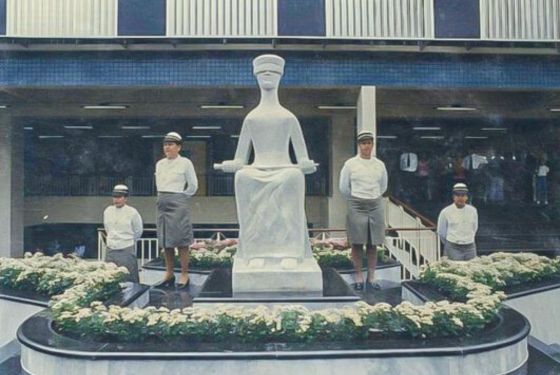 Foto da estátua da justiça em frente ao Fórum Barão do Rio Branco. Ao lado da obra, tem quatro PMs mulheres, duas de cada lado posando para foto.