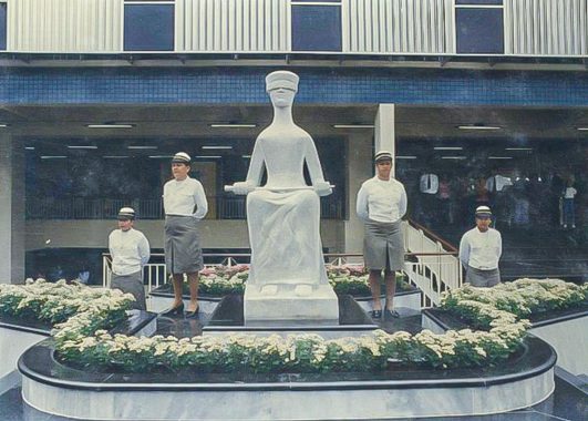 Foto da deusa Themis no dia da reinauguração do Fórum Barão de Rio Branco. Ao seu redor há flores brancas plantadas em um pequeno jardim e quatro mulheres militares, disposta duas de cada lado