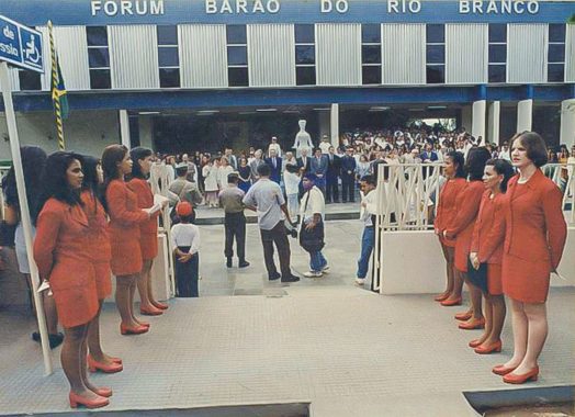 Foto da solenidade de na reinauguração do Fórum Barão de Rio Branco. As pessoas do cerimonial estão em fileiradas nos dois lados da entrada do fórum. A frente há muitas pessoas aguardando o evento.