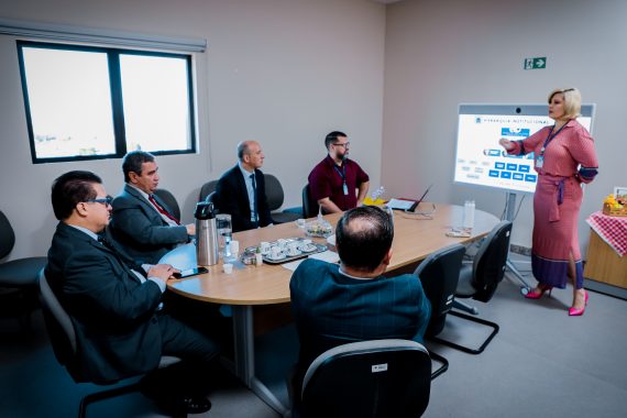 Foto de uma mesa de reunião, onde um servidor e o desembargador Élcio Mendes assistem uma explanação. Há uma mulher em pé explicando um gráfico que está projetado na parede