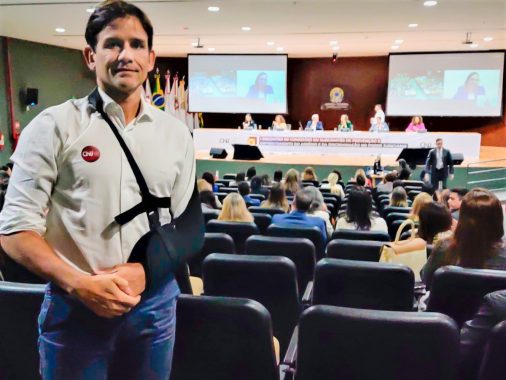 Foto do juiz Danniel Bomfim no auditório onde está ocorrendo o encontro sobre assédio. Ele está em primeiro plano, o braço está em uma tipóia e na sua camisa tem um adesivo do CNJ. Ao fundo está a mesa de debates e os participantes assistindo ao evento.