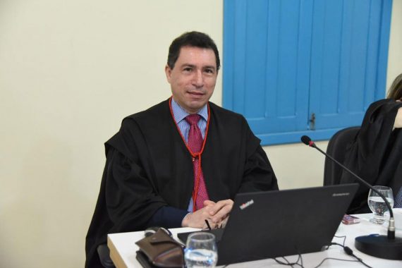 Foto do desembargador Laudivon Nogueira. Ele está sentado com as mãos entrelaçadas, sorrindo para foto. Há um notebook e um copo de água à sua frente. Ele está usando toga e está no Centro Cultural do Juruá.