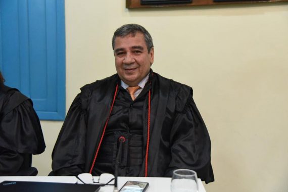 Foto do desembargador Élcio Mendes. Ele está usando toga, sorrindo para foto e o óculos está na mesa à sua frente. Ele também está no Centro Cultural do Juruá.