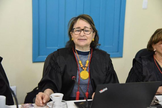 Foto da desembargadora Denise Bonfim. Ela está sentada, com um notebook a sua frente. Há uma xícara de café. Ela está usando um colar amarelo e a toga. Ela está no Centro Cultural do Juruá.