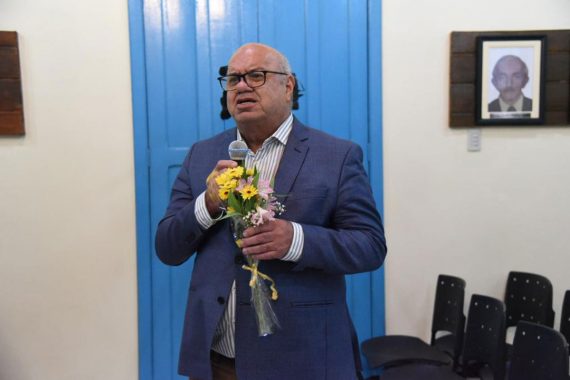 Foto do desembargador Arquilau de Castro falando ao microfone no Centro Cultural do Juruá e segurando flores amarelas.