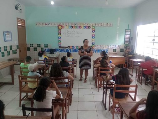 Foto da sala de aula Escola Frei André Maria Fiarelli. A professora está a frente do quadro explicando e a sala está cheia de crianças em suas carteiras.
