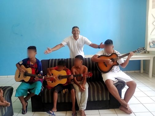 Foto do voluntário com braços abertos e quatro crianças estão sentadas no sofá tendo aula de música