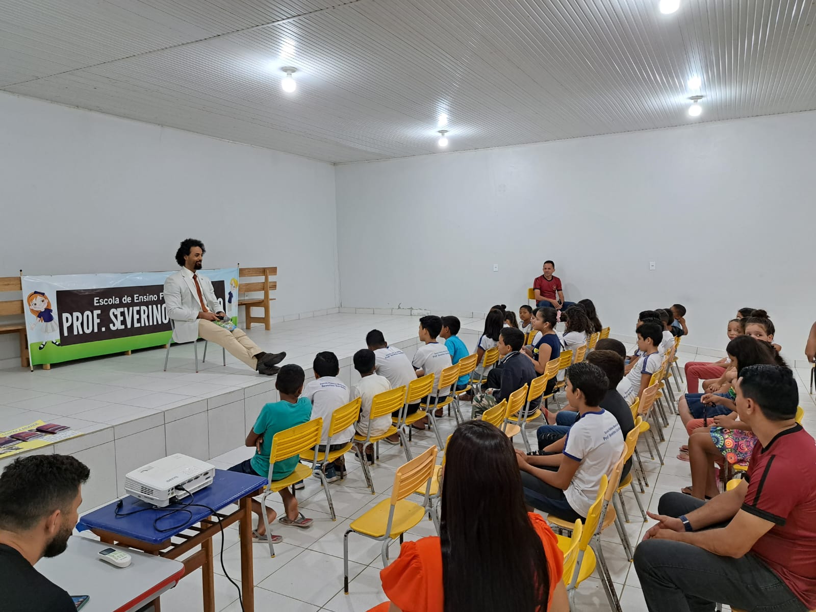 Na foto o Promotor de Justiça, Lucas Nonato canto superior esquerdo de vestimenta branca mediando conversa com as crianças sobre o Poder Judiciário, atrás do Promotor de Justiça, um cartaz escrito Escola de Ensino Municipal Prof. Severino Cordeiro