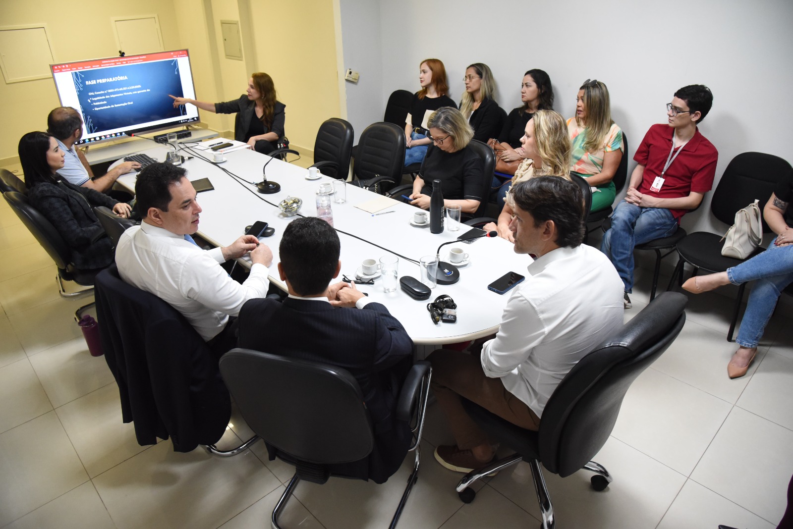 Mulher, possivelmente juíza de Direito, aponta para uma apresentação na televisão. Na foto há a presença 9 mulheres e 5 homens, todos estão sentados em uma sala de paredes brancas.