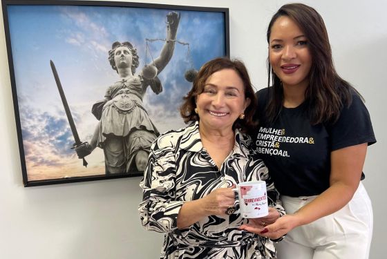 Em frente a um quadro com uma foto da deusa Themis, símbolo da Justiça, estão a desembargadora Eva Evangelista sorrindo e segurando uma caneca que ganhou da pessoa que está ao seu lado, a produtora do documentário "Sobreviventes", Marcy Bezerra. A caneca é uma divulgação da obra audiovisual.