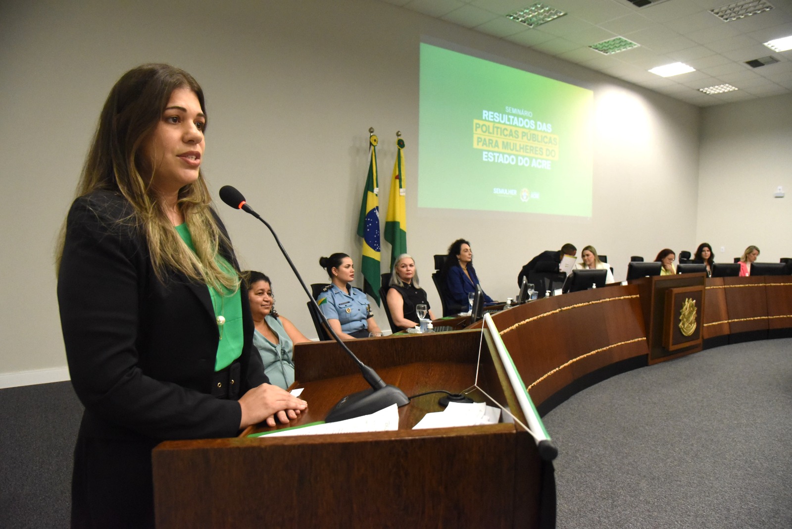 Imagem da Juíza Louise Kristina no palanque. Ao seu lado esquerdo, há oito mulheres sentadas, todas autoridades participantes da solenidade.