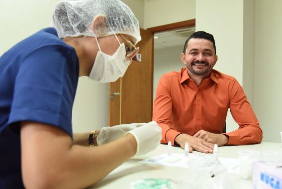 A imagem mostra um ambiente clínico onde um profissional da saúde, vestindo jaleco azul, máscara, touca e luvas, está realizando um procedimento, possivelmente um teste rápido. Ela manipula pequenos frascos e dispositivos de teste sobre uma mesa. À frente dela, sentado e sorridente, está um homem de camisa social laranja, observando o processo com expressão tranquila e amigável. Ele aparenta estar sendo atendido pela profissional. Sobre a mesa há copos descartáveis, frascos com reagentes, materiais de teste e uma caixa com a marca “NUGARD”, possivelmente de luvas descartáveis. A cena transmite um clima de cuidado, atenção e acolhimento, comum em ações de saúde preventiva, como campanhas de exames rápidos ou triagens em ambientes institucionais.