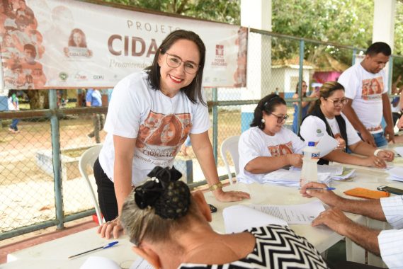 imagem mostra um momento de atendimento no Projeto Cidadão, uma iniciativa que promove o acesso à justiça e à cidadania. A mulher ao centro, vestindo a camisa do projeto, sorri enquanto atende uma senhora que está assinando documentos. Na mesa, há papéis, canetas e garrafas de água, sugerindo um ambiente de trabalho voltado ao auxílio comunitário. O Projeto Cidadão busca oferecer serviços essenciais, como emissão de documentos, registro civil e orientação jurídica, facilitando o acesso a direitos básicos para a população. O cenário ao ar livre, cercado por árvores e uma quadra esportiva, indica que as ações acontecem em espaços comunitários, reforçando a proximidade com a sociedade. Esse tipo de iniciativa tem um impacto significativo, pois garante que pessoas em situação de vulnerabilidade tenham seus direitos reconhecidos e formalizados. O sorriso da atendente reflete o espírito do projeto: acolhimento, empatia e compromisso com a cidadania.