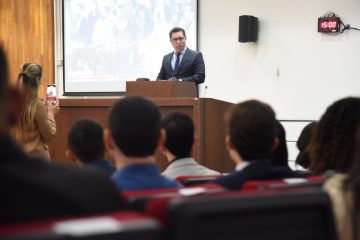 A imagem mostra uma cerimônia ou evento formal acontecendo em um auditório. Um homem de terno escuro e gravata está em destaque, discursando em um púlpito de madeira no palco. Atrás dele, há uma tela de projeção com uma imagem exibida, embora não esteja completamente visível. A parede do fundo é revestida com painéis de madeira clara, o que dá um aspecto institucional ao ambiente. O público está atento, sentado em poltronas vermelhas, com algumas pessoas de pé ou em movimento na lateral do auditório. Duas pessoas próximas ao palco estão tirando fotos ou gravando vídeos com celulares, indicando que o momento é significativo, possivelmente a fala de uma autoridade ou entrega de certificados. No alto da parede lateral, há um relógio digital marcando 15:00, o que reforça o ambiente organizado e controlado do evento. A plateia é composta por adultos de várias idades, vestidos de maneira formal ou semiformal, em clima de concentração e respeito.