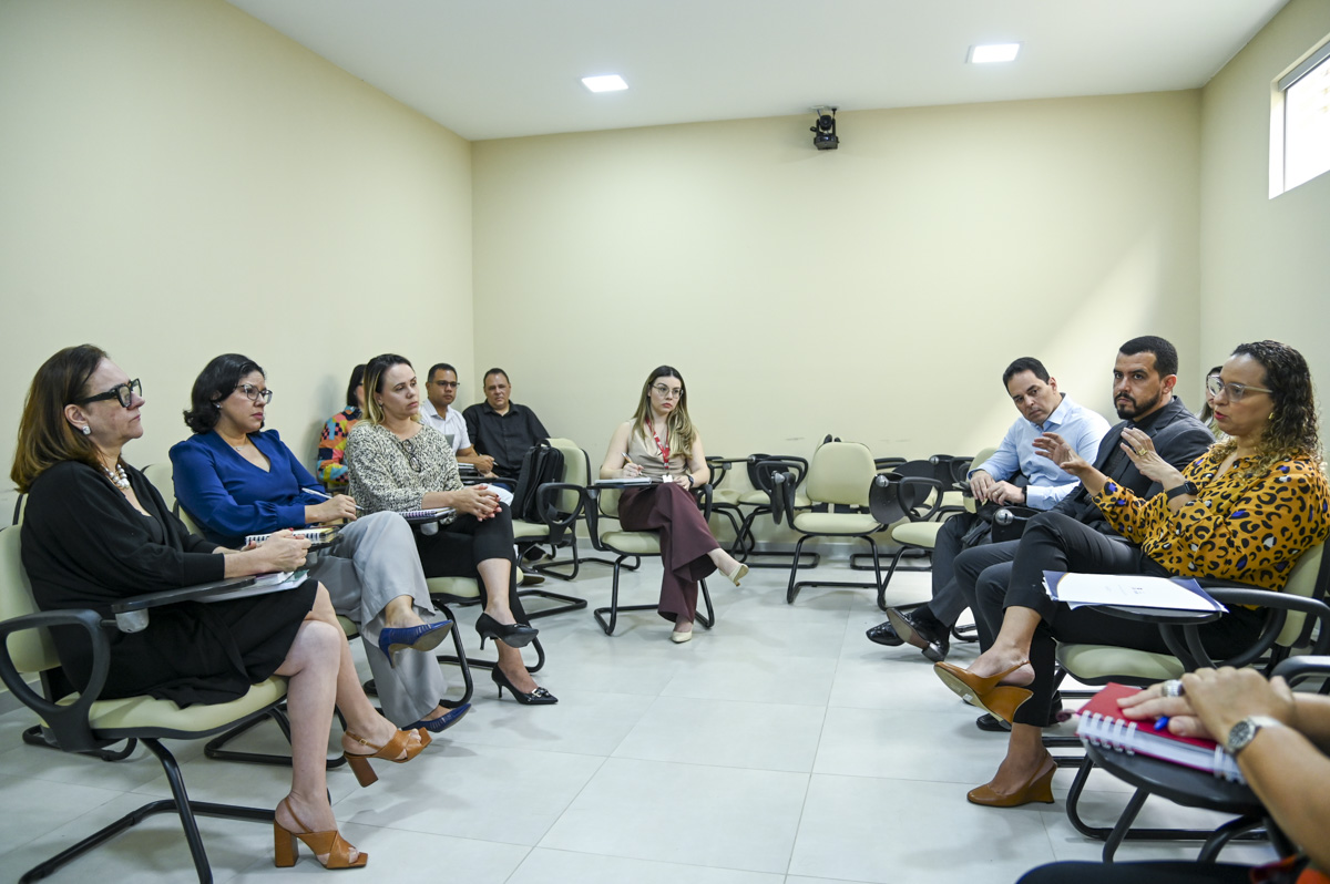 A imagem mostra uma reunião presencial em andamento em uma sala de conferências moderna. O ambiente é iluminado, com paredes claras e piso de cerâmica clara, criando um espaço limpo e organizado. As pessoas estão dispostas em círculo, sentadas em cadeiras com apoio de braço e prancheta acoplada, sugerindo um formato de discussão ou apresentação colaborativa. Detalhes observados: Participantes: Aproximadamente 11 pessoas participam da reunião, sendo homens e mulheres de diferentes idades. Todos estão bem-vestidos, com roupas formais ou sociais, indicando um contexto corporativo ou institucional. Várias pessoas estão com cadernos ou blocos de anotações no colo, demonstrando atenção e engajamento. Uma mulher do lado direito (de vestido preto e óculos) parece estar falando, gesticulando enquanto se dirige ao grupo — possivelmente liderando ou apresentando algo. Ambiente: Um monitor grande está fixado na parede ao fundo, exibindo o que parece ser uma apresentação ou uma ferramenta de gestão com gráficos e texto (difícil de ler com clareza na imagem). Há uma caixa de som na parede superior esquerda e um aparelho de ar-condicionado na mesma parede. Ao fundo, vê-se uma mesa com garrafa térmica e copos, indicando a disponibilidade de café ou água para os participantes. A cena transmite um momento de alinhamento estratégico, planejamento ou apresentação de resultados.