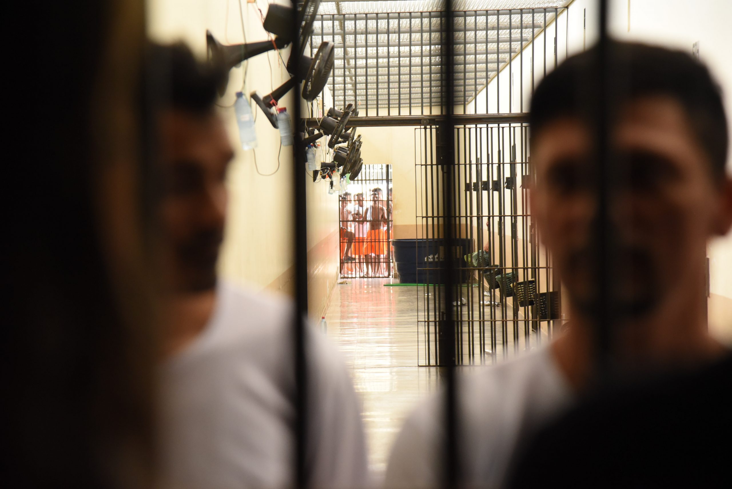 Uma imagem mostra um corredor de prisão com grades, onde vários detentos estão presos. A frente se encontra duas pessoas com camisa brancas em desfoco.