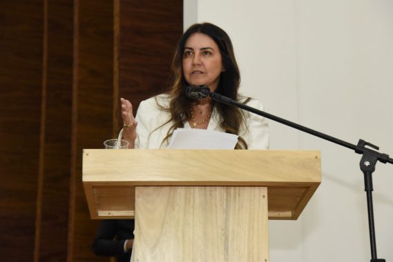 A imagem mostra uma mulher fazendo um discurso ou apresentação em um púlpito de madeira claro. Ela está vestida com um blazer branco e segura alguns papéis na mão esquerda, enquanto gesticula com a mão direita, transmitindo expressividade em sua fala. Há um copo com água sobre o púlpito, e um microfone direcionado para sua voz está posicionado em um suporte articulado. O fundo é dividido entre uma parede branca e uma parede com painéis de madeira escura, criando um ambiente formal e institucional, possivelmente relacionado a uma cerimônia, conferência ou evento público. A mulher aparenta estar concentrada e determinada, com uma postura firme e segura. O cenário reforça uma atmosfera de seriedade e profissionalismo.