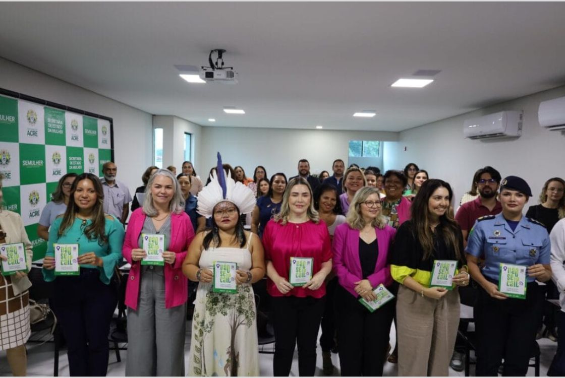 A imagem mostra um grupo diverso de pessoas reunido em um auditório durante um evento institucional, aparentemente promovido pela Secretaria de Estado da Mulher do Acre (SEMULHER), conforme indicado pelo painel de fundo. O evento parece estar relacionado ao combate à violência contra a mulher ou à promoção de direitos femininos, já que várias mulheres na primeira fileira seguram um livreto com o título “Quebre o Silêncio”. Na linha de frente, estão mulheres de diferentes perfis: indígenas, servidoras públicas, profissionais uniformizadas (incluindo uma policial militar), e outras representantes da sociedade civil. A presença de uma mulher com cocar indica representatividade dos povos indígenas no evento. Ao fundo, mais participantes assistem de pé, demonstrando envolvimento com a causa. O ambiente é moderno e bem iluminado, com um projetor no teto e ar-condicionado nas paredes, sugerindo uma estrutura institucional organizada. O clima da imagem transmite engajamento e união em prol de uma causa social relevante.