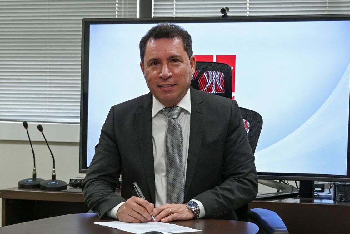 A imagem mostra o homem assinando um documento ou contrato sobre o púlpito e possando pra foto. O homem veste terno preto com a gravata cinza. A composição e o ambiente indicam uma ocasião formal, possivelmente uma cerimônia de entrega ou reconhecimento institucional.