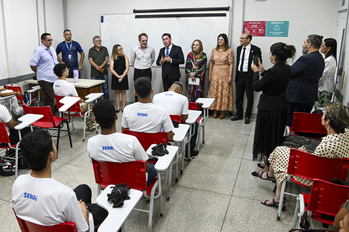 A imagem mostra um momento formal em uma sala de aula do SENAI, onde um grupo de autoridades e gestores está de pé à frente de uma lousa, se dirigindo a um grupo de alunos sentados. Os estudantes vestem uniformes brancos com a marca "SENAI" e estão atentos ao que está sendo dito. As cadeiras vermelhas e o ambiente limpo e organizado reforçam o ambiente educacional. Ao centro, um homem de terno e gravata azul parece estar discursando ou fazendo uma apresentação. Ao seu lado, estão outros quatro adultos — dois homens e duas mulheres — trajando roupas formais. O cenário sugere uma visita institucional, anúncio de parceria, inauguração ou entrega de equipamentos, reforçando o papel do SENAI na qualificação profissional.