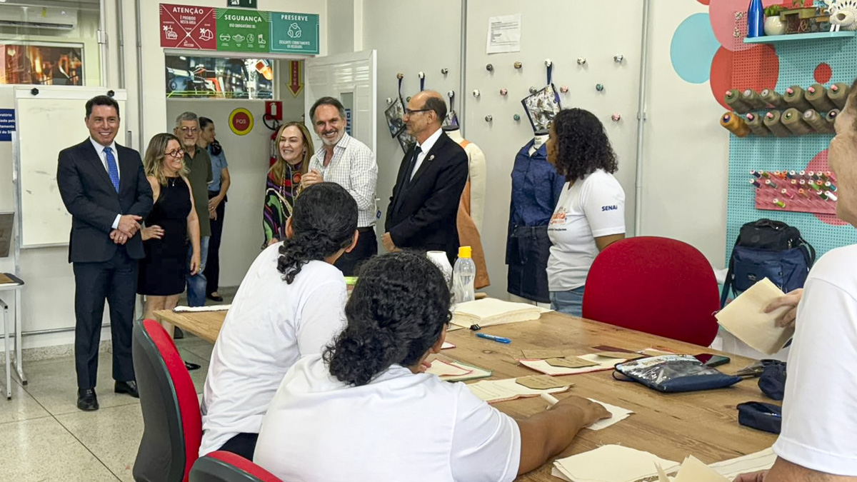 A imagem mostra uma visita institucional a uma sala de aula ou laboratório de costura, possivelmente dentro de uma unidade do SENAI (Serviço Nacional de Aprendizagem Industrial), como indicado nas camisetas de alguns presentes. Na parte esquerda da imagem, um grupo de pessoas — incluindo homens de terno e uma mulher com vestido colorido — observa e interage com os alunos. Eles aparentam ser autoridades ou visitantes importantes. Os alunos, usando camisetas brancas com o logotipo do SENAI, estão sentados ao redor de uma mesa grande de madeira, trabalhando com moldes de costura ou peças de tecido. O ambiente é colorido e organizado, com carretéis de linha e aventais pendurados na parede ao fundo. Há sinalizações de segurança e orientação na entrada da sala, reforçando um ambiente de aprendizagem técnica e profissional.