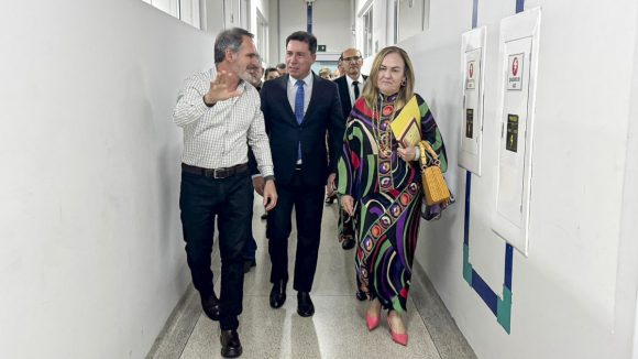 Três pessoas estão paradas, em poses formais, em frente a uma parede clara. A composição da imagem é bastante simples, com os três indivíduos dispostos em uma linha horizontal, posicionados de forma simétrica, mas não em linha reta, frente a uma parede branca. O foco principal parece estar nas três pessoas, com a parede como pano de fundo neutro. Não há elementos visuais adicionais que distram do grupo. Os três indivíduos são adultos. O homem à esquerda usa uma camisa de gingham e o homem ao centro, um terno escuro. A mulher, à direita, veste um casaco colorido. As expressões faciais são neutras, sem emoções fortes visíveis. O homem à esquerda parece manter as mãos juntas em forma de saudação. As roupas parecem apropriadas para um evento formal ou reunião. O estilo da imagem é fotográfico, com iluminação e foco aparentemente direcionados aos indivíduos. A qualidade da imagem é boa, sem distorções visíveis ou efeitos especiais. A composição é bem equilibrada, sem elementos deslocados. A iluminação é uniforme, mas não excessivamente brilhante, permitindo ver os detalhes do rosto e das roupas de cada pessoa. A atmosfera é de profissionalismo e formalidade. O ambiente parece ser um evento ou reunião, como uma conferência ou coletiva de imprensa, devido às roupas e poses. A iluminação controlada e as posições formais sugerem um evento estruturado.