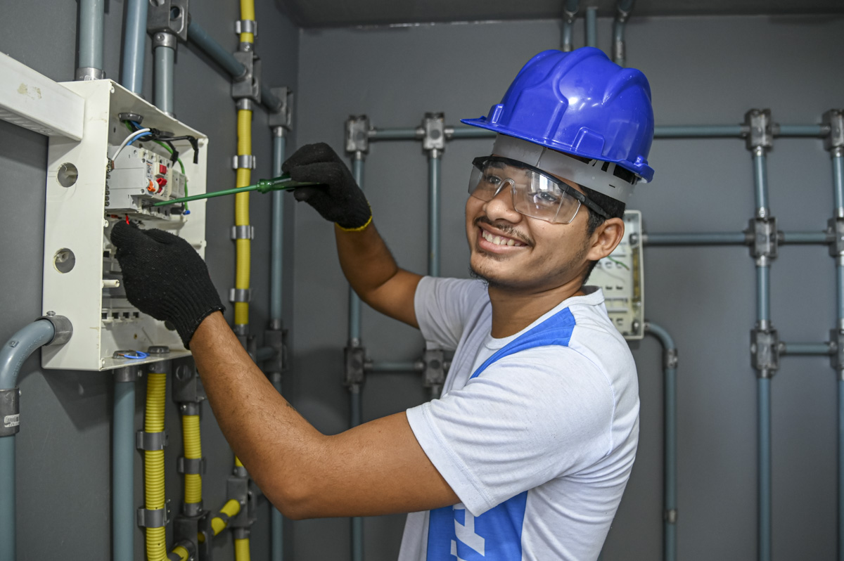 Um jovem trabalhador realiza reparos em uma caixa de distribuição elétrica. A composição da imagem apresenta um trabalhador de perfil, posicionado de forma inclinada para a direita, em frente a uma caixa de distribuição elétrica. Ele está concentrado em um trabalho de manutenção ou reparo. A postura do indivíduo demonstra foco e atenção na tarefa. O enquadramento da imagem é fechado, concentrando-se no trabalhador e no seu trabalho. O sujeito principal é um jovem, possivelmente adolescente, de aparência amistosa. Ele usa roupas de trabalho, incluindo uma camiseta branca com detalhes azuis, luvas pretas e óculos de segurança. Um capacete azul claro complementa seu uniforme. Seu rosto e seus olhos demonstram concentração, e ele tem um olhar calmo e atencioso. O meio técnico é fotográfico, apresentando uma qualidade de imagem nítida e focada, com boa iluminação que realça as características do sujeito. Não há estilização artística ou efeitos gráficos visíveis. O cenário é interno, em um ambiente que parece ser um local de trabalho ou oficina. O fundo apresenta tubos metálicos e conexões, sugerindo uma estrutura elétrica. As paredes são cinzas, criando um fundo discreto e focado no trabalho. A iluminação é adequada, sem sombras fortes ou destaques excessivos, proporcionando um ambiente de trabalho bem iluminado. A atmosfera é de trabalho e profissionalismo.