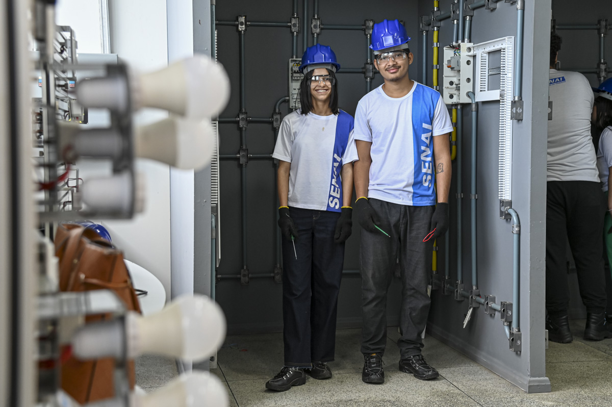 A imagem mostra dois jovens sorridentes, usando capacetes azuis, óculos de proteção e luvas de segurança, em um ambiente de treinamento técnico. Ambos vestem uniformes com a logomarca do SENAI (Serviço Nacional de Aprendizagem Industrial), indicando que são alunos ou participantes de um curso técnico, provavelmente na área de eletricidade ou eletrotécnica. Eles estão posicionados em frente a um painel didático de instalações elétricas, com tubulações, tomadas, interruptores e fios visíveis. À esquerda da imagem, há um painel com lâmpadas e, ao fundo, mais alunos também uniformizados interagindo com os equipamentos. A atmosfera da imagem é positiva e demonstra um ambiente educacional prático, voltado à formação profissional.