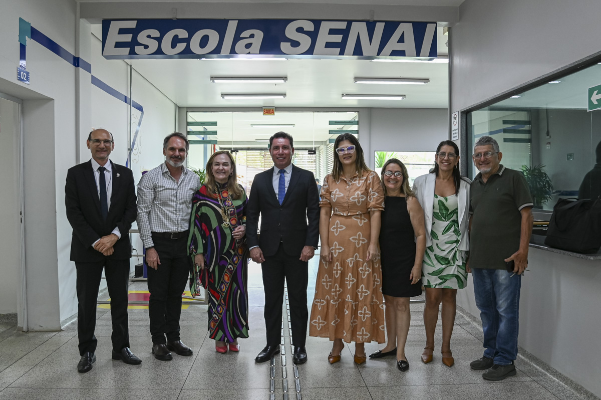 A imagem mostra um grupo de oito pessoas posando para uma foto em frente a uma entrada com uma faixa azul escrita “Escola SENAI”. O ambiente parece ser interno, com piso de granito e iluminação fluorescente. As pessoas estão bem vestidas, indicando que se trata de um evento formal ou visita institucional. Ao fundo, vê-se uma porta de vidro e sinalizações como “saída” e uma seta verde indicando direção. O grupo é composto por homens e mulheres, a maioria sorrindo para a câmera. À direita da imagem, há um balcão com um espelho ou vidro refletindo parte do ambiente.