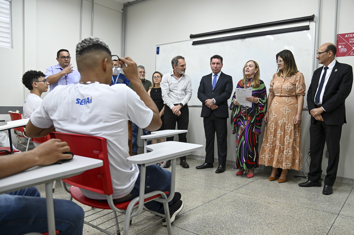 A imagem mostra sete pessoas em pé atras uma parede cor branco, e um jovem sentado mexendo no celular. Os homens vestem ternos a combinar cor preto, as mulheres vestidos cores marrom claro com detalhe uma delas, a outra veste com detalhe de listra coloridas e sapatos a combinar, a outra mulher um vestido preto a combina os sapatos. O ambiente transmite uma sala corporativa e escola com estudantes uniformizados na sala.