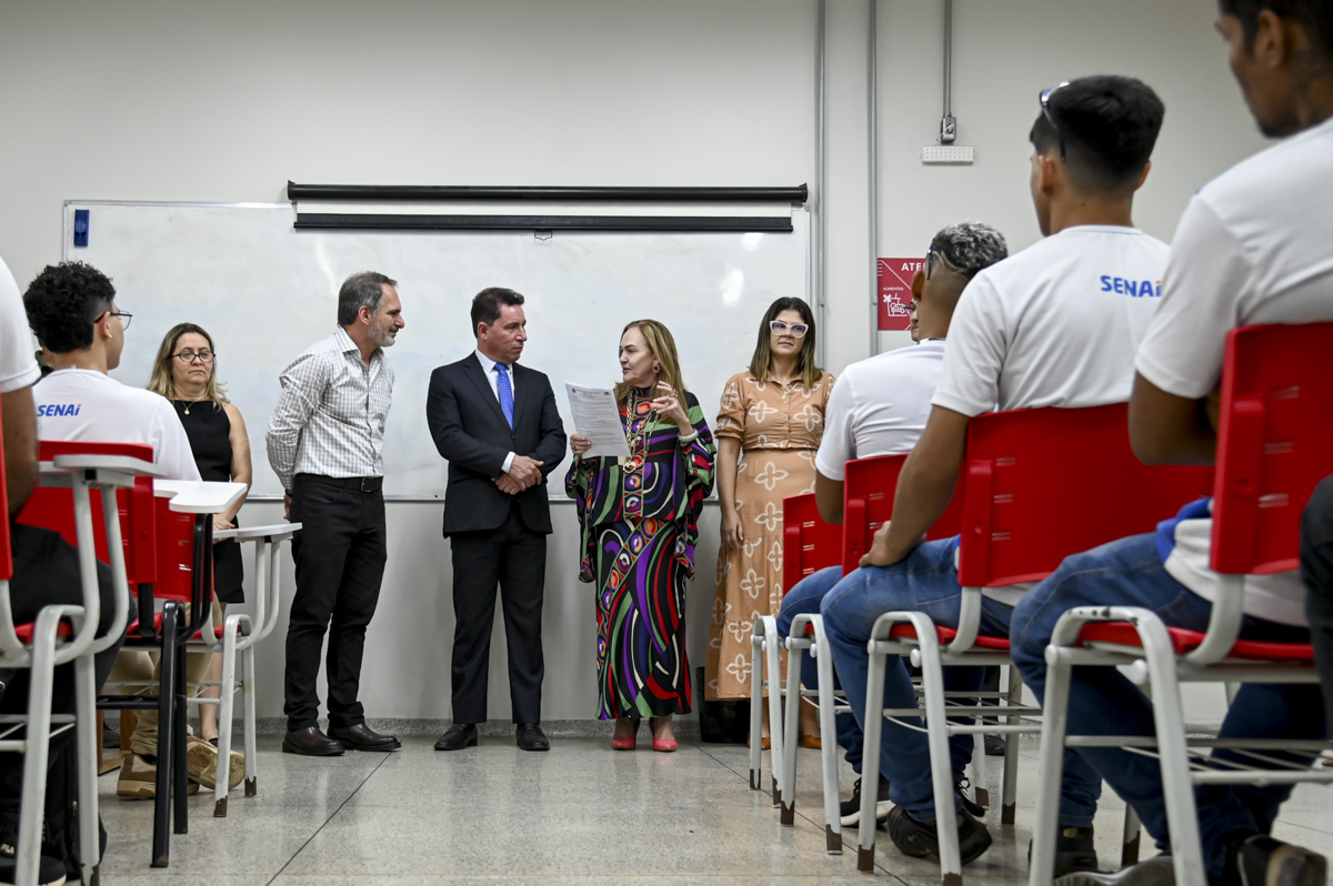 A imagem mostra sete pessoas em pé atras uma parede cor branco, e um jovem sentado mexendo no celular. Os homens vestem ternos a combinar cor preto, as mulheres vestidos cores marrom claro com detalhe uma delas, a outra veste com detalhe de listra coloridas e sapatos a combinar, a outra mulher um vestido preto a combina os sapatos. O ambiente transmite uma sala corporativa e escola com estudantes uniformizados na sala.