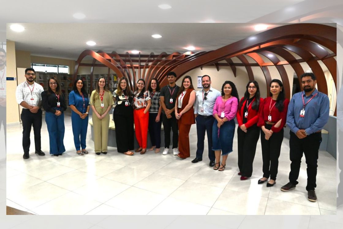 A imagem mostra um grupo de quinze pessoas (seis homens e nove mulheres) posando para uma foto em um ambiente interno bem iluminado e moderno. Eles estão alinhados lado a lado, formando uma fileira diante de uma estrutura arquitetônica decorativa de madeira curvada, que lembra uma escultura ou instalação artística. Todos usam crachás vermelhos no pescoço, o que indica que podem ser funcionários de uma mesma instituição, como uma empresa, órgão público ou universidade. Estão vestidos de maneira formal ou semiformal, sugerindo um ambiente corporativo ou institucional. Ao fundo, há prateleiras com livros ou documentos, o que pode indicar que a foto foi tirada em uma biblioteca, centro de documentação ou espaço administrativo. O clima da imagem é profissional e cordial.