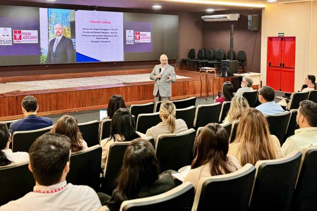 A imagem mostra uma palestra sendo realizada em um auditório. Um homem de terno cinza claro, identificado no telão como Eduardo Sabbag, está em pé no centro do espaço, falando para o público. No fundo, há uma grande tela com informações sobre ele, indicando que é: Doutor em Língua Portuguesa (PUC-SP) Doutor em Direito Tributário (PUC-SP) Professor universitário (Mackenzie/SP e Famerp/AM) Advogado tributarista O telão também exibe os logotipos da ESJUD (Escola do Poder Judiciário do Estado do Acre) e do Poder Judiciário do Estado do Acre, sugerindo que o evento é promovido por essa instituição. O público, composto por homens e mulheres sentados em poltronas pretas, assiste atentamente à apresentação. O ambiente é bem iluminado, com portas vermelhas ao fundo e um palco elevado de madeira. A atmosfera é formal e acadêmica, voltada à capacitação ou atualização profissional.