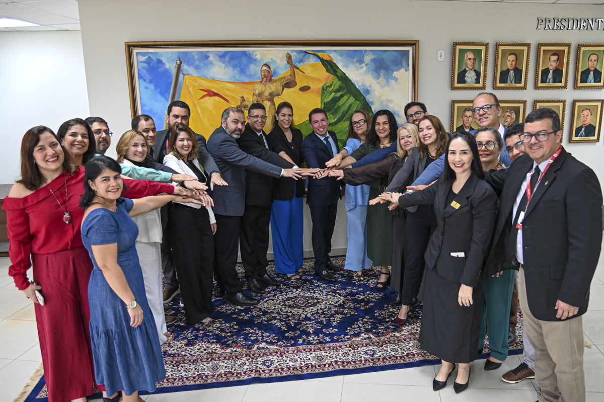 Em ambiente institucional, grupo de gestores do Tribunal de Justiça do Acre reúne-se em semicírculo com o desembargador-presidente, em trajes formais, com as mãos estendidas ao centro em gesto de união. Ao fundo, há um quadro artístico e retratos oficiais, compondo o cenário solene do encontro.